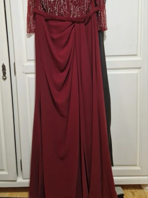 Vinröd långklänning med paljetter - Elegant vinröd långklänning med paljettprydd överdel och långa ärmar. Kjolen har draperade detaljer och ett bälte i midjan. Perfekt för speciella tillfällen där du vill sticka ut med en glamorös look.