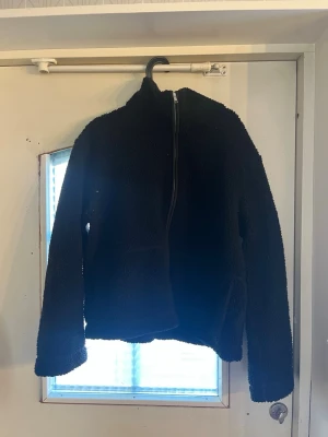 Svart teddyjacka med dragkedja - Mysig svart teddyjacka med hög krage och hel dragkedja framtill. Jackan har två stora fickor på framsidan och är tillverkad i fluffigt teddy-material som håller dig varm. Perfekt för kalla dagar och enkel att matcha med olika outfits.
