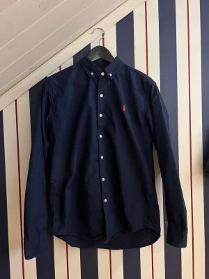 Mörkblå skjorta från Ralph Lauren - Klassisk mörkblå skjorta från Ralph Lauren med button-down krage och lång ärm. Skjortan har vita knappar och den ikoniska röda broderade loggan på bröstet. Tillverkad i bomull för en bekväm och stilren look.