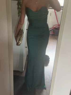 Grön axelbandslös långklänning - Elegant grön långklänning med figurnära passform och axelbandslös design. Klänningen har draperad överdel och markerad midja som framhäver siluetten. Perf brudtärna eller bal. Materialet ser ut att vara mjukt och följsamt, troligen polyester. Aldrig använd 