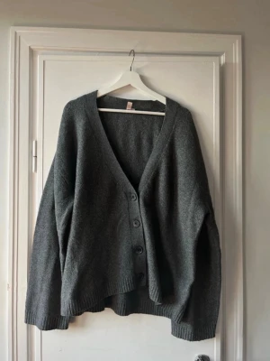 Mörkgrå oversized kofta med knappar - Mysig och oversized mörkgrå kofta med djup V-ringning och fyra knappar framtill. Koftan har långa ärmar och ribbade muddar vid ärmslut och nederkant. Perfekt för lager-på-lager och chill stil.