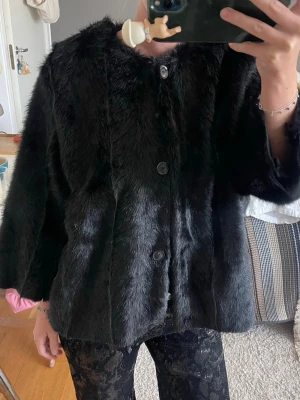 Svart pälsjacka med knappar - Snygg svart pälsjacka med rak passform och stora knappar framtill. Jackan har en klassisk, lite boxig siluett och är tillverkad i mjukt fuskpälsmaterial. Perfekt för att hålla värmen under kalla dagar och ger en lyxig känsla till din outfit.