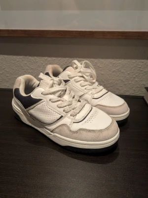Vita sneakers med grå och svarta detaljer - Snygga vita sneakers med grå mockadetaljer och svarta partier vid hälen. Skorna har perforerade partier på sidorna och framtill för extra ventilation. Klassisk snörning och låg profil gör dem perfekta till streetwear-looken.