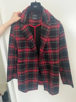 Rutig kavaj från Bershka - Snygg kavaj i svart och rött rutmönster från Breshka. Dubbelknäppt modell med klassisk krage och två framfickor. Perfekt för dig som vill sticka ut med en trendig och färgstark look. Materialet känns mjukt och följsamt.