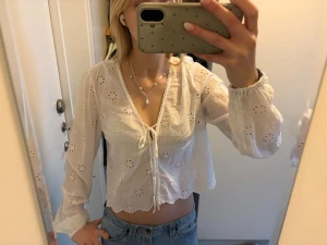 Vit broderad blus med knytning - Söt vit blus med broderade blommor och knytning framtill. Blusen har v-ringning, långa ärmar med resår vid handlederna och vågig nederkant. Tunt, luftigt material som ger en romantisk känsla.