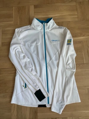 Vit träningsjacka från Craft - Snygg vit träningsjacka från Craft med blå detaljer och hel dragkedja. Jackan har hög krage, långärmad design och svensk flagga på ena ärmen. Perfekt för sport och utomhusaktiviteter. Materialet är funktionellt och lätt, idealiskt för träning.