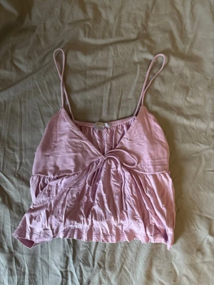 Ljusrosa crop top med tunna band - Söt ljusrosa crop top med tunna axelband och knytning framtill. Toppen har en luftig och avslappnad passform med lätt rynkade detaljer. Perfekt för sommardagar och enkel att matcha med jeans eller kjol.