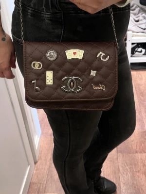 Brun handväska med kedjerem - Säljer en brun quiltad handväska  med silverfärgade detaljer och dekorativa spelmotiv på locket. Väskan har en klassisk kedjerem och ikonisk CC-logga framtill. Perfekt för dig som gillar unika accessoarer och lyxig design.