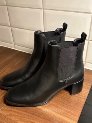 Svarta Chelsea boots med klack från Skopunkten - Stilrena svarta Chelsea boots i slätt skinn med elastiska paneler på sidorna och dragflikar både fram och bak. Bootsens klack är bred och stabil, vilket ger en cool siluett. Perfekta till jeans eller kjol för en trendig look.