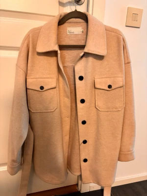 Beige kappa från NLY Trend - Stilren beige tunn kappa från NLY Trend med stora bröstfickor, svarta knappar och bälte i midjan. Den har en rak passform, klassisk krage och är tillverkad i ett mjukt material som ger en avslappnad look.