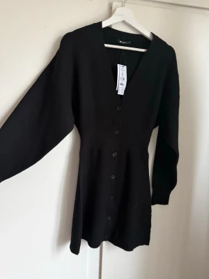 Svart stickad klänning från Gina Tricot - Svart stickad klänning från Gina Tricot med v-ringning och knappar längs hela framsidan. Klänningen har lång ärm och markerad midja, Storlek xs, aldrig använd