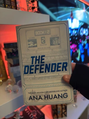 The Defender av Ana Huang - FIRST EDTION. Säljer boken 'The Defender' av Ana Huang, bok två i serien 'Gods of the Game'. Omslaget är beige med blå text och sporttema, och boken är i pocketformat. Perfekt för dig som gillar romantisk skönlitteratur med sportinslag.