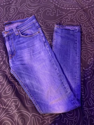 Blå slim fit jeans från Nudie Jeans 32/32 - Säljerz ett par blå slim fit jeans från Nudie Jeans med klassisk femficksmodell och snygga slitningar. Jeansen har en vågig detalj på bakfickorna och är tillverkade i mjuk denim. Perfekta för dig som gillar en avslappnad men stilren look.
