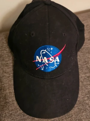 Svart NASA keps med broderad logga - Svart keps med klassisk böjd skärm och broderad NASA-logga i blått, vitt och rött framtill. Kepsen har ventilerande hål och justerbar baksida.  