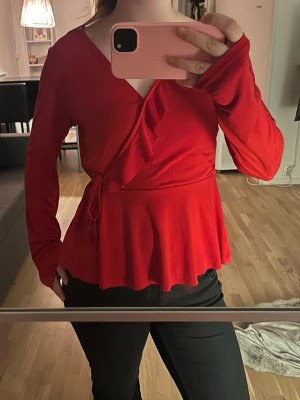 Röd omlottblus med volang - Trendig röd omlottblus med långa ärmar och volangdetalj framtill. Knytband i midjan för en snygg passform och lätt utsvängd nederkant. Perfekt att styla med både jeans och kjol för en chic look.
