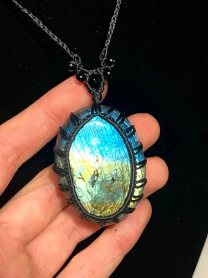 Talisman med labradorit - Unikt halsband med en stor, oval labradorit i blått och gult, infattad i svart makramé. Labradorit sägs vara en sten för intuition, skydd och inre styrka. Den skimrande ytan symboliserar dolda möjligheter och hjälper bäraren att följa sin egen väg. En talisman som många använder för att stärka sin energi och sitt inre lugn.