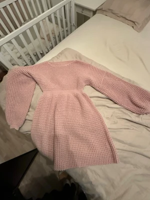 Rosa stickad klänning med lång ärm - Mysig rosa stickad klänning med lång ärm och markerad midja. Klänningen har en kortare längd och ett grovt stickat mönster som ger en avslappnad men ändå söt look. Perfekt för kyligare dagar och enkel att styla med boots eller sneakers.
