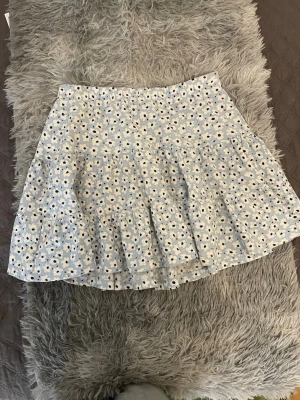 Blommig volangkjol i ljusblått - Söt kortkjol med volanger i ljusblått tyg med små vita och svarta blommor. Kjolen har en avslappnad passform och är perfekt för vår och sommar. Ingen synlig dragkedja eller knappar, bara ett mjukt och flowy material.