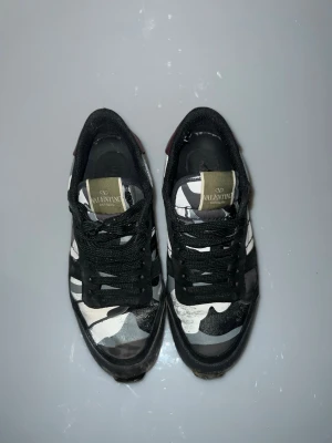 Valentino rockrunners i camo - Snygga sneakers från Valentino med svart och vit camo-mönster, svarta skosnören och detaljer i vinrött på hälen. Ovandelen är i textil och mocka, med gummisula för skön komfort. Perfekta för dig som vill sticka ut med en unik stil. Till behör inget boxen gick tyvärr sönder 