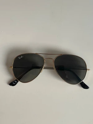 Ray-Ban aviator solglasögon guld - Klassiska Ray-Ban aviator solglasögon med tunna guldfärgade metallbågar och mörka linser. Snygg tidlös design med svarta detaljer på skalmarna. Perfekta för dig som vill ha en ikonisk och stilren look.