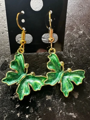 Gröna fjärilsörhängen i metall - Unika örhängen formade som fjärilar i skimrande grönt med guldfärgade detaljer. Hängande modell med krok, tillverkade i metall och emalj. Perfekta för dig som vill sticka ut med ett lekfullt och färgstarkt smycke.
