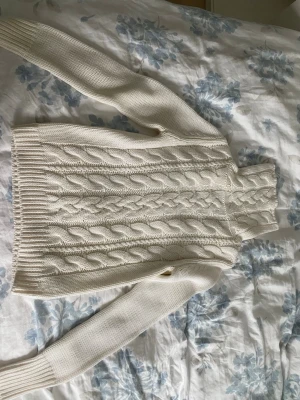 Vit kabelstickad polotröja - Säljer en vit kabelstickad polotröja med höga ribbade muddar och lång ärm. Tröjan har ett klassiskt flätmönster framtill och är gjord i mjukt stickat material. Perfekt för att hålla sig varm och snygg under kyliga dagar. Finns fler bilder att få!