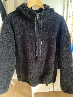 Svart teddyjacka med huva - Mysig svart teddyjacka med huva och hel dragkedja framtill. Jackan har en bröstficka med dragkedja och justerbar nederkant med snören. Perfekt för kalla dagar och ger en avslappnad look. Materialet är fluffigt och mjukt, vilket gör den extra bekväm.