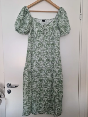 Gina Tricot grön blommig klänning med puffärm - Söt grön klänning med vitt blommönster och puffiga korta ärmar. Klänningen har v-ringning, liten rosett framtill och en slits nedtill. Perfekt för sommarens alla tillfällen och har en luftig passform. Storlek L, normal i storleken
