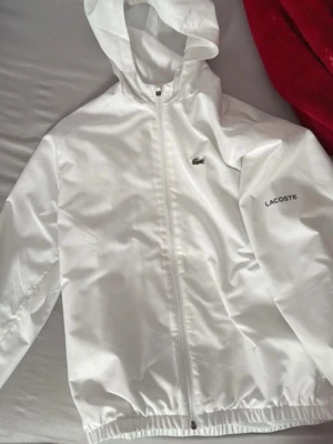 Vit vindjacka med huva från Lacoste - Snygg vit vindjacka från Lacoste med dragkedja framtill och huva. Jackan har elastisk nederkant och ärmar, samt diskret Lacoste-logga på bröstet. Tillverkad i lätt och vindtätt material, perfekt för blåsiga dagar.