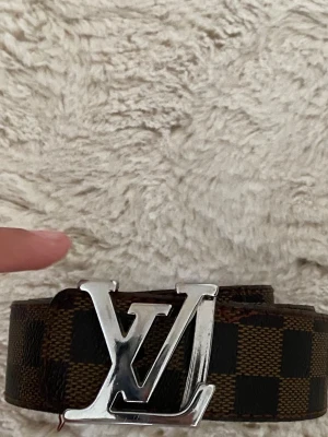 Brunt rutigt bälte i skinn - Snyggt brunt bälte  med klassiskt rutigt mönster och stor silverfärgad logga som spänne. Bältet är gjort i skinn och har en exklusiv känsla som lyfter vilken outfit som helst. Fick den som present och använde den inte längre 