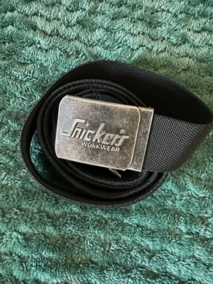 Svart bälte från Snickers Workwear - Snyggt svart bälte från Snickers Workwear med en robust metallspänne där loggan är ingraverad. Bältet är gjort i slitstarkt tygmaterial och har en klassisk, enkel design som passar till de flesta outfits.