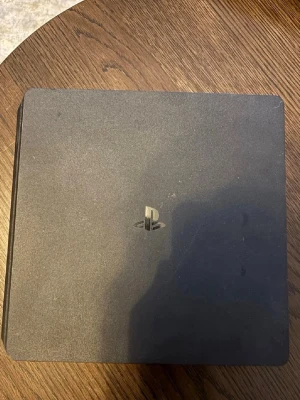 PlayStation 4 Slim med handkontroll - Säljer en svart PlayStation 4 Slim från Sony. Konsolen har en stilren, rektangulär design med rundade hörn och PlayStation-loggan på ovansidan. Materialet är matt plast. HDMI-kabel medföljer för enkel anslutning till TV eller skärm.