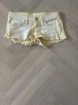 Ljusgula jeansshorts med fransig kant - Säljer ett par ljusgula jeansshorts med fransig, rå kant och klassisk femficksdesign. Shortsen har låg midja och knappgylf. Perfekta för varma dagar och ger en avslappnad vibe.