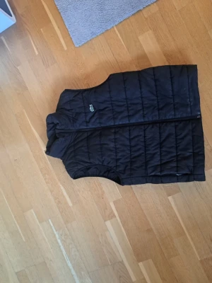 Svart  dunväst från Lacoste - Snygg svart dunväst från Lacoste med klassisk ståkrage och diskret logga på bröstet. Västen är quiltad och har dragkedja framtill. Perfekt att slänga över en hoodie eller tröja för extra värme under kyliga dagar.