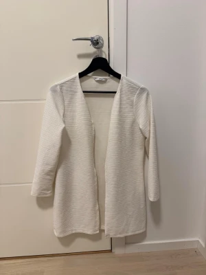 Vit cardiganjacka från ONLY - Stilren vit kappa/kofta  från ONLY i öppen modell utan knappar. Kappan har en rak passform, trekvartsärmar och är tillverkad i ett mjukt, ribbat tyg. Perfekt för lager på lager och enkel att matcha med olika outfits.