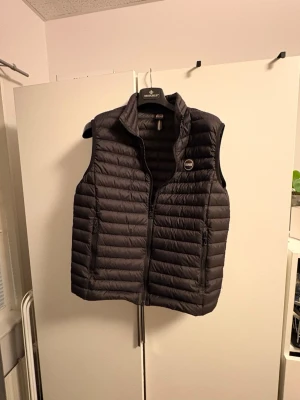 Svart dunväst från colmar i storlek m - Säljer en svart dunväst från Colmar med quiltad design och dragkedja framtill. Västen har två sidofickor med dragkedja och en hög krage. Perfekt att ha över en hoodie för extra värme. Märkets logga sitter på bröstet. Hör av er vid funderingar 