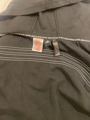 Svart Adidas hoodie med dragkedja - Säljer en svart hoodie från Adidas med dragkedja framtill och klassiska vita ränder längs ärmarna. Adidas-logga på bröstet och huva. Tillverkad i mjukt bomullsmaterial och har textdetaljer på insidan. Perfekt för en sportig och avslappnad stil.