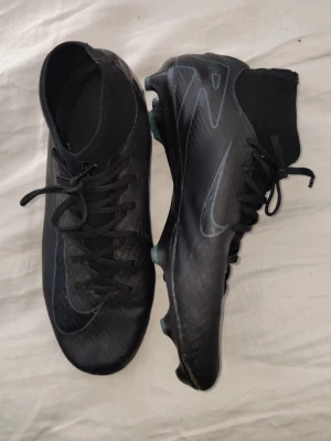 Svarta Nike fotbollsskor med högt skaft - Snygga svarta fotbollsskor från Nike med högt skaft och snörning. Skorna har en stilren design med diskret Nike-logga på sidan och är tillverkade i syntetmaterial för bra grepp och känsla på planen. Perfekta för dig som vill ha kontroll och stil på fotbollsplanen.