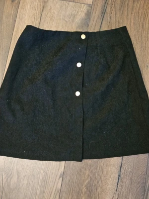 Svart kjol med knappar framtill - En svart kortkjol med tre silverfärgade knappar framtill. Kjolen har en enkel och stilren design med lätt A-linjeformad siluett. Perfekt att matcha med både stickade tröjor och t-shirts.