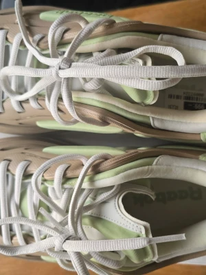 Reebok sneakers i pastellgrönt och beige - Snygga Reebok sneakers med pastellgröna och beige detaljer, vita skosnören och en chunky sula. Ovandelen är i textil och syntetmaterial, och sulan har ett coolt rutmönster undertill. Perfekta för dig som gillar en sportig och trendig look.