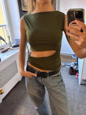 Grön croppad topp med slits - Säljer en mörkgrön croppad topp med cool slitsdetalj vid midjan. Toppen är ärmlös och har en tight passform som framhäver figuren. Perfekt för dig som gillar trendiga och unika plagg. Materialet känns som syntet och är stretchigt.