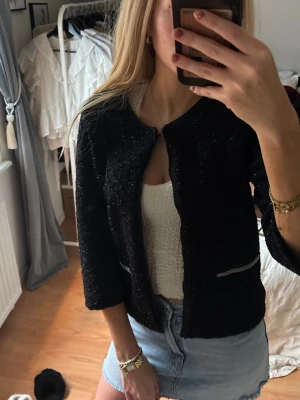 Svart glittrig cardigan med fejk dragkedjor - Snygg svart kofta med diskret glittrigt tyg och trekvartsärmar. Jackan har rund halsringning och två dekorativa dragkedjor framtill. Perfekt att slänga över en topp för en trendig look.