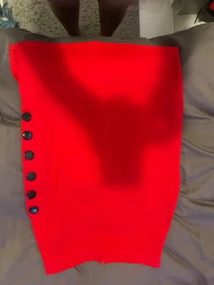 Röd kjol med knappar i sidan - Snygg röd kjol med sex svarta knappar längs ena sidan. Kjolen har en rak, något figurnära passform och är gjord i ett stretchigt material som sitter bekvämt. Perfekt för dig som vill sticka ut med en färgstark look.