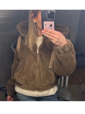 Brun teddyjacka med huva - Mysig brun teddyjacka med dragkedja framtill och stor huva. Jackan har en avslappnad passform, resår vid ärmslut och midja samt mjukt, fluffigt material. Perfekt för kalla dagar när du vill vara både varm och stylish.