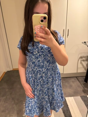 Blå blommig kortklänning - Säljer en söt blå kortklänning med vita blommor. Klänningen har korta ärmar och en rund halsringning. Den är lätt och luftig, perfekt för varma dagar. 