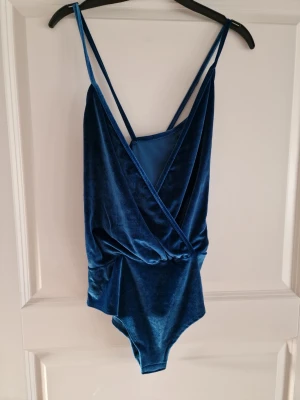 Sammetsbody med omlott - Supersnygg blå body i mjuk sammet från H&M. Modellen har smala axelband som korsas i ryggen och en omlottringning framtill. Perfekt för dig som vill ha något trendigt och bekvämt. Materialet är stretchigt och följsamt för en skön passform.