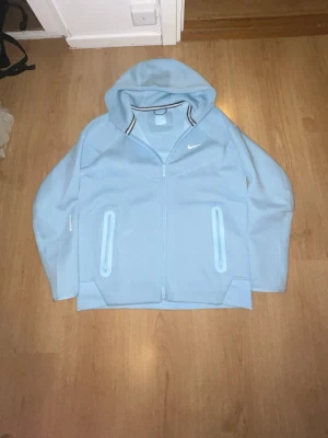 Ljusblå Nike NOCTA hoodie - Snygg ljusblå hoodie från Nike NOCTA med dragkedja framtill och två fickor. Hoodien har en vit Nike-logga på bröstet och coola detaljer vid huvan. Tillverkad i mjukt material som känns skönt mot huden. Perfekt för en avslappnad streetwear-look.