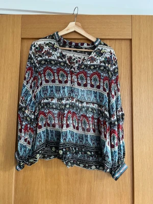 Mönstrad blus från Zara - Färgglad blus från Zara med bohemiskt mönster i rött, blått, svart och vitt. Blusen har rund halsringning, knappar framtill och långa ärmar med resår vid ärmslut. Tunt, luftigt material som ger en avslappnad och trendig look.