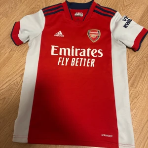 Arsenal röd och vit fotbollströja Adidas - Säljer en officiell Arsenal fotbollströja från Adidas med klassisk röd front och vita sidor och ärmar. Tröjan har korta ärmar, klubbmärke på bröstet och Emirates Fly Better-tryck. Materialet är lätt och andas, perfekt för match eller träning.