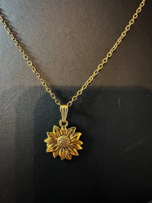 Guldigt halsband med solrosberlock - Snyggt halsband i guldfärgad metall med en detaljrik solros som hänge. Kedjan är tunn och har små ovala länkar som ger ett elegant intryck. Perfekt för dig som gillar blommiga smycken och vill ha något som sticker ut lite extra.